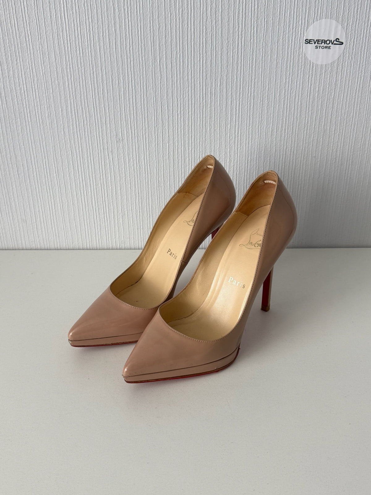 A pair of pointed-toe high heels in a light nude beige patent leather with signature red soles, displayed on a light gray surface against a textured gray wall.